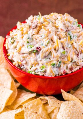 Side view of a bright red bowl filled high with prepared million dollar dip, pictured surrounded by tortilla chips.