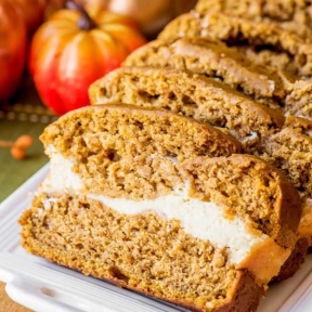 Side view of slices of prepared cream cheese pumpkin bread laying on a white plate, pictured on a fall-themed cloth with pumpkins in the background.