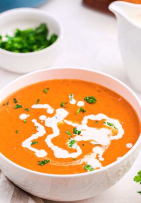 Side view of a bowl full of tomato bisque with heavy cream and parsley as garnish.