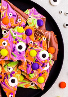 Close up top down view of multiple pieces of Halloween chocolate bark on a black plate, surrounded by more candy eyes and M&M's.