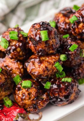 Side view of a long white plate piled high with prepared and sauced Asian chicken meatballs, garnished with chopped green onion and pictured with a cooked red chili pepper.