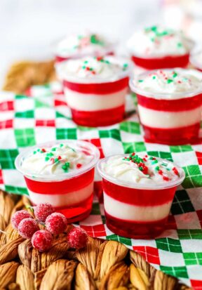 Side view of multiple prepared and decorated candy cane jello shots resting on a woven mat and decorative Christmas-themed cloth.