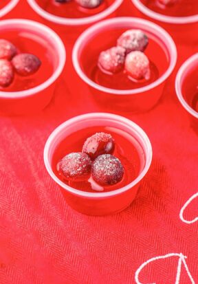 Side view of multiple vibrant red cranberry jello shots placed on a red holiday cloth.