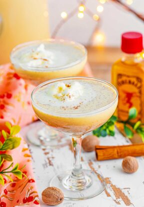 Side view of two cocktail glasses filled with eggnog punch, pictured on a wooden board surrounded by garland, cinnamon sticks, walnuts, Christmas lights, and bottles of Fireball Whisky.