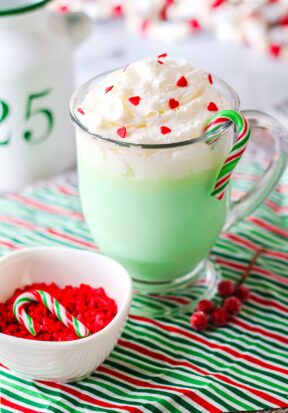 Side view of a prepared Grinch hot chocolate, decorated with whipped cream and heart sprinkles, pictured with a bowl of heart sprinkles.