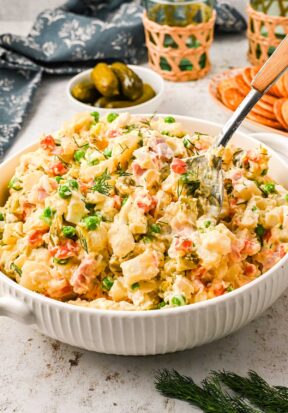 Side view of a large bowl filled with ham potato salad, pictured with a spoon dug in the middle and surrounded by more ingredients like fresh dill, dill pickles, carrots, etc.