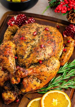 Close up side view of a freshly baked orange herb roasted chicken, pictured with fresh rosemary, orange slices, and pomegranate seeds.