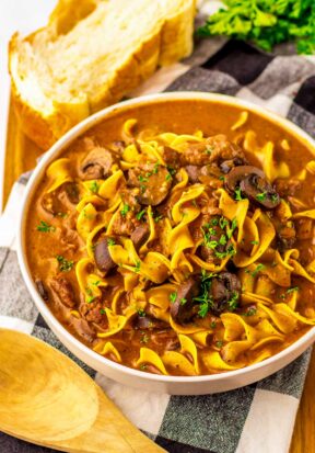 Top down side view of a large bowl filled with prepared slow cooker beef stroganoff, garnished with parsley and pictured with crispy bread.