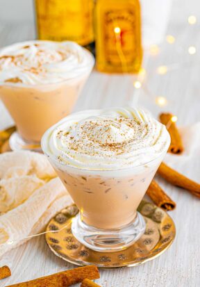 Side view of two prepared cinnamon roll cocktails in festive glasses, pictured with cinnamon sticks and twinkle lights as garnished.