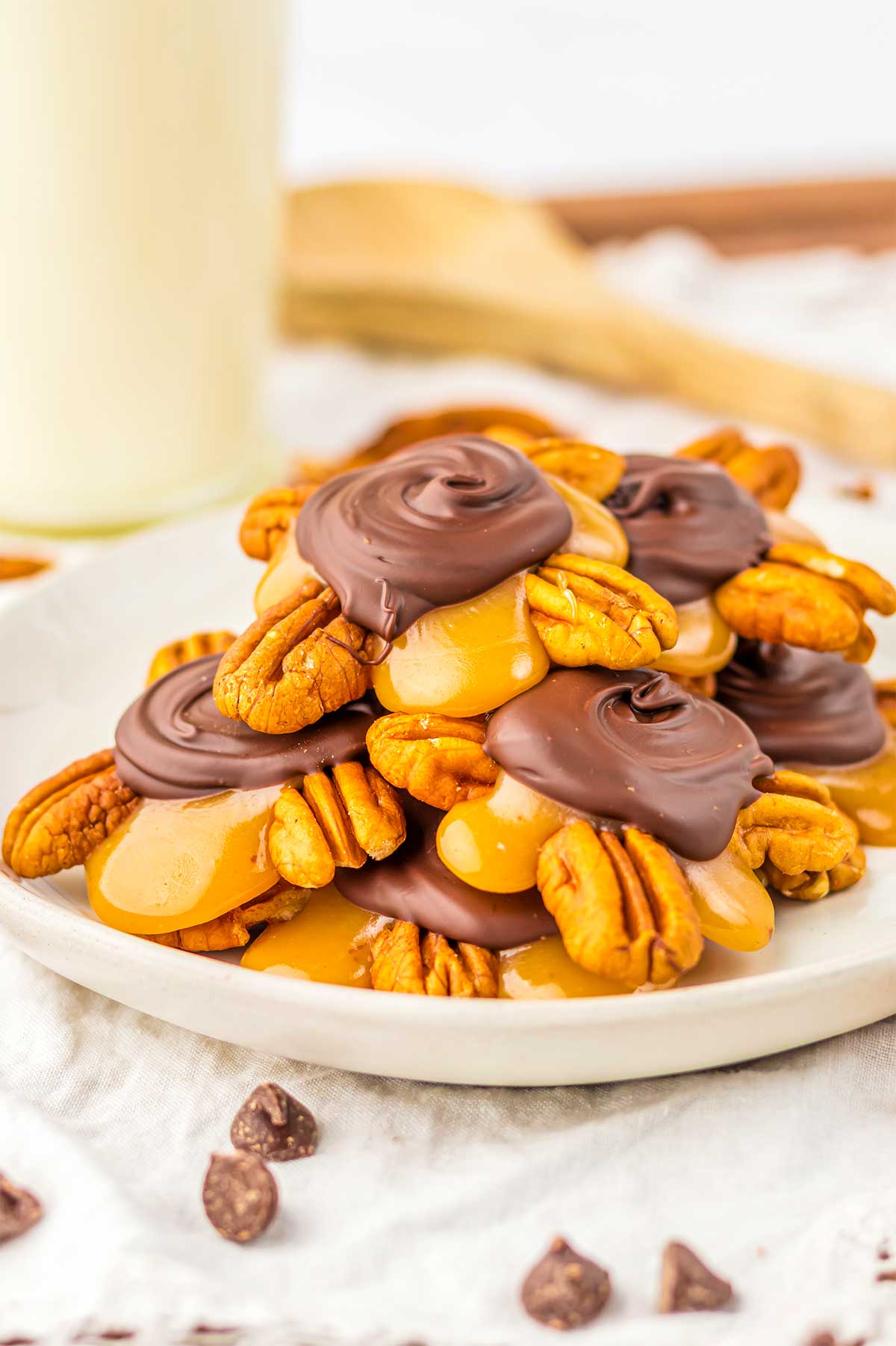 Side view of a plate piled high with prepared turtle candies, pictured surrounded with chocolate chips and pecans.