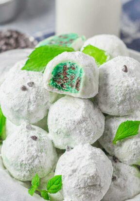 Side view of a bowl piled high with powdered sugar-covered peppermint snowball cookies, garnished with a few bright green mint leaves, pictured with the top most cookie with a bite taken out, showing off the green filling and laced with mini chocolate chips.