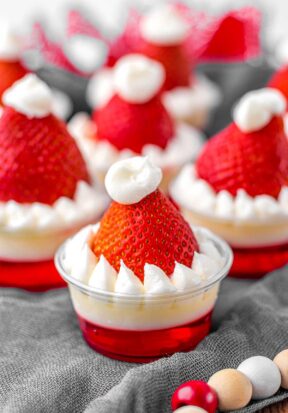 Side view of multiple prepared Santa hat jello shots resting on a gray cloth, surrounded by beaded garland.