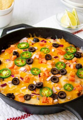 Side view of a freshly prepared baked taco dip, pictured in a black cast iron skillet and topped with fresh jalapeno slices, black olives, Roma tomatoes, and red onion.