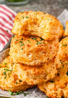 Close up side view of a pile of cheddar bay biscuits, focusing on 3 in the front, stacked, with the top most biscuit with a bite taken out, showing off the flaky interior.