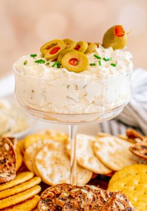 Side view of dirty martini dip served in a flat bottomed martini glass, garnished with olives and chives, pictured on a wooden tray topped with many different options of crisp crackers for dipping.