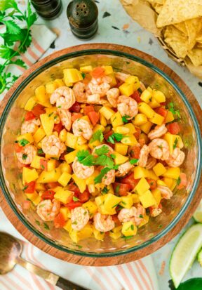 Top down view of a bowl filled with freshly made mango shrimp salsa, garnished with extra cilantro on top, and pictured surrounded with fresh ingredients like peppers, limes, tortilla chips, and bunches of cilantro.