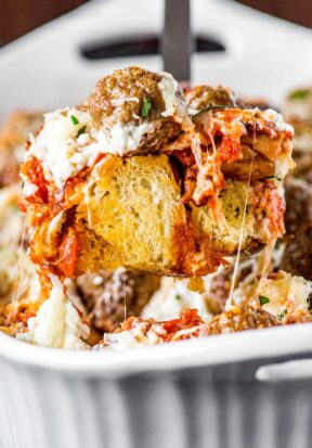 Close up side view of a spatula lifting up a serving of meatball sub casserole, showing off the layers of garlic bread, cheese, meatballs, and sauce, with delicious strings of cheese pulling down.