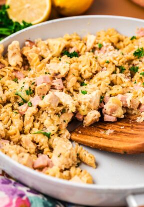 Close up side view of a skillet filled with freshly prepared chicken cordon bleu pasta, pictured with a wooden spatula dug under some of the pasta, ready to life it up and serve.