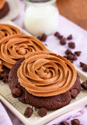 Close up side view of multiple chocolate cake cookies pictured lined up on a narrow plate, surrounded by more chocolate chips and a glass of milk.