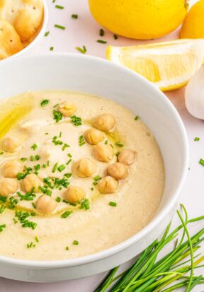 Side view of a bowl filled with garlic hummus, garnished with whole chickpeas, chopped parsley, and olive oil, and pictured surrounded by whole chives, toasted bread, and lemon slices.