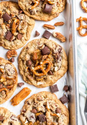 Top down view of freshly baked kitchen sink cookies still on the baking sheet, surrounded by more toppings like soft caramels, chocolate chunks, and pretzels.