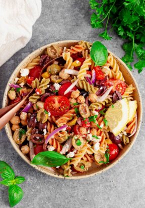 Close up top down view of prepared Mediterranean pasta salad, pictured in a pale-colored bowl with a white cloth and more fresh herbs sprinkled around, with two spoons dug into the salad, ready to serve.