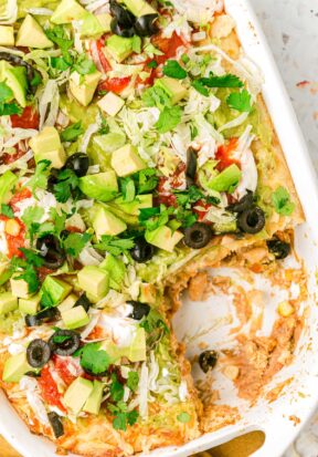 Top down view of prepared and garnished white chicken taco casserole, focusing on a corner area that's had a square piece of casserole already served.