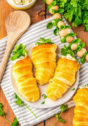 Top down view of a plate with 3 crescent roll carrots on it, pictured with more parsley garnish, a bowl of chicken salad, and a wooden spoon.