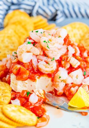 Side view of a serving plate topped with cream cheese shrimp dip, pictured surrounded by Ritz crackers, and a small spreading knife that's resting on the plate, having already dug into the dip, showing off the vibrant red sauce shrimp and the bright white cream cheese underneath.