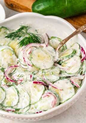 Side view of a bowl filled with creamy cucumber salad, pictured with an antique spoon dug into it, ready to serve.