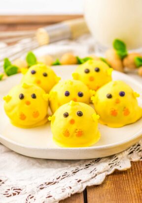 Side view of multiple Easter chick truffles arranged on a white plate, all facing the camera, pictured with wooden garnish and a glass of milk in the background.