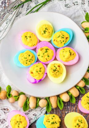 Top down view of a plate covered with multiple Easter deviled eggs with pink, purple, yellow, and blue egg whites, surrounded by a wooden garnish and pictured with more deviled eggs placed around.