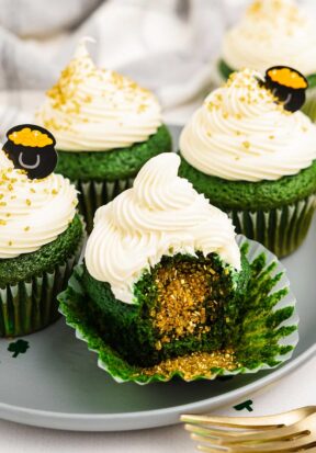 Side view of multiple cupcakes on a plate, pictured with the center most cupcake with the wrapper rolled down and a bite taken out, showing off the golden sprinkles spilling out from inside.