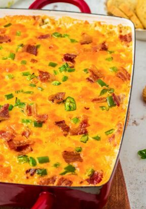 Close up side view of a deep casserole dished filled to the top of freshly baked jalapeno popper chicken casserole, showing off the cheesy top covered with sliced jalapenos, crumbled bacon, and green onions.